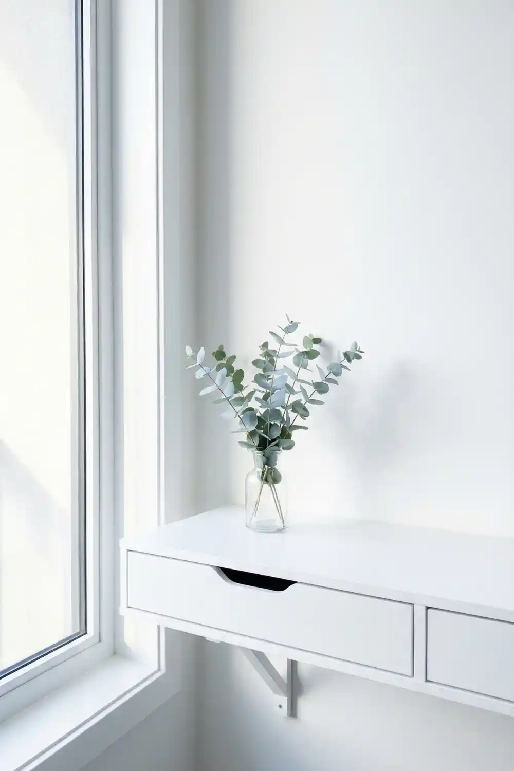 Minimal indoor plant decor with fresh eucalyptus stems in clear glass vase on white floating desk