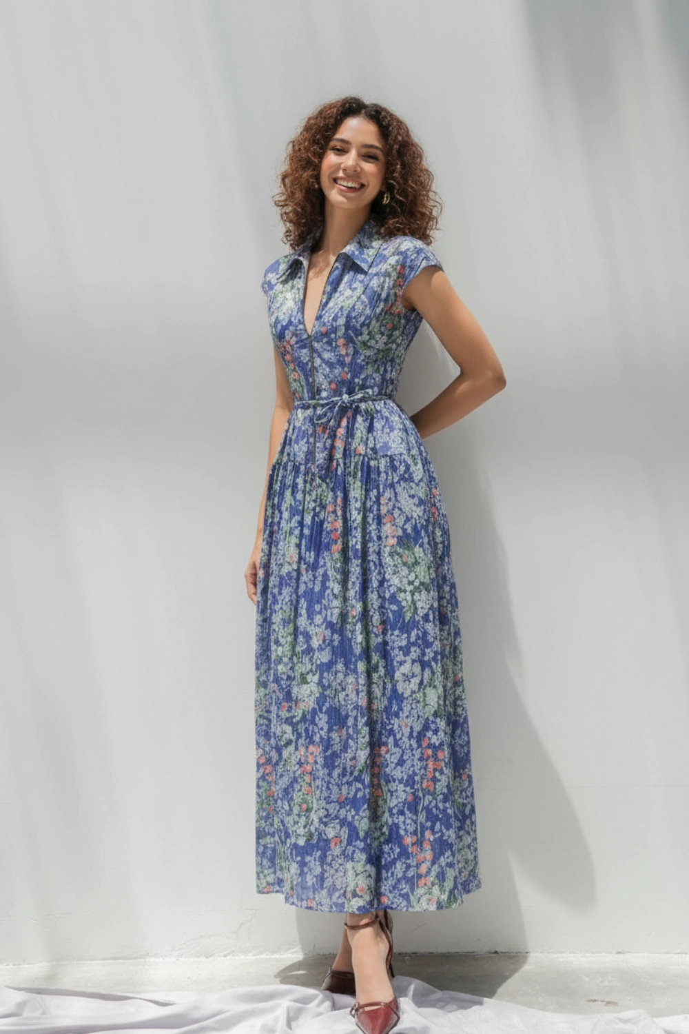Woman wearing a blue floral dress with a belted waist and maroon heels against a white wall