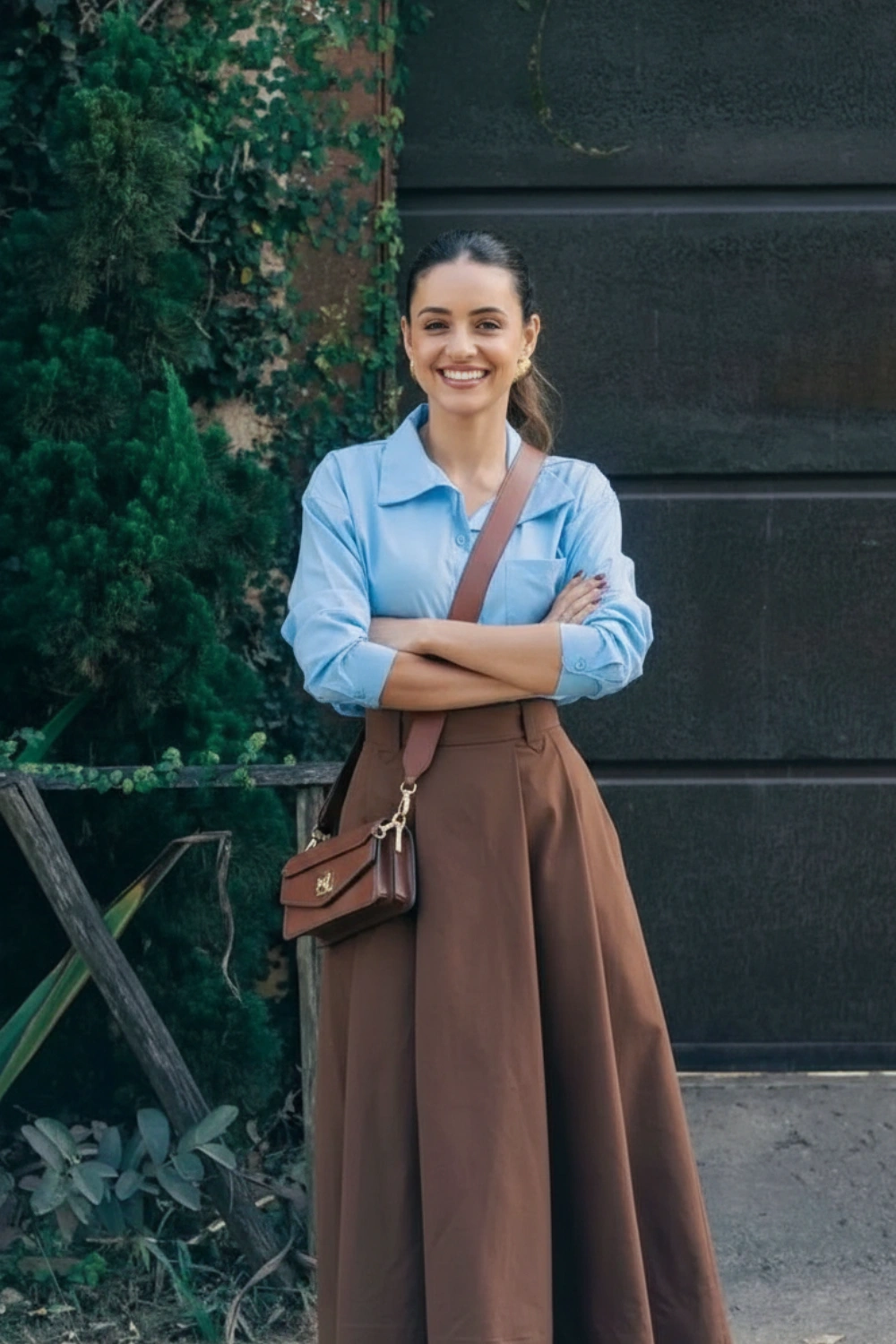 Woman wearing a light blue shirt with a brown skirt and brown crossbody bag outdoors