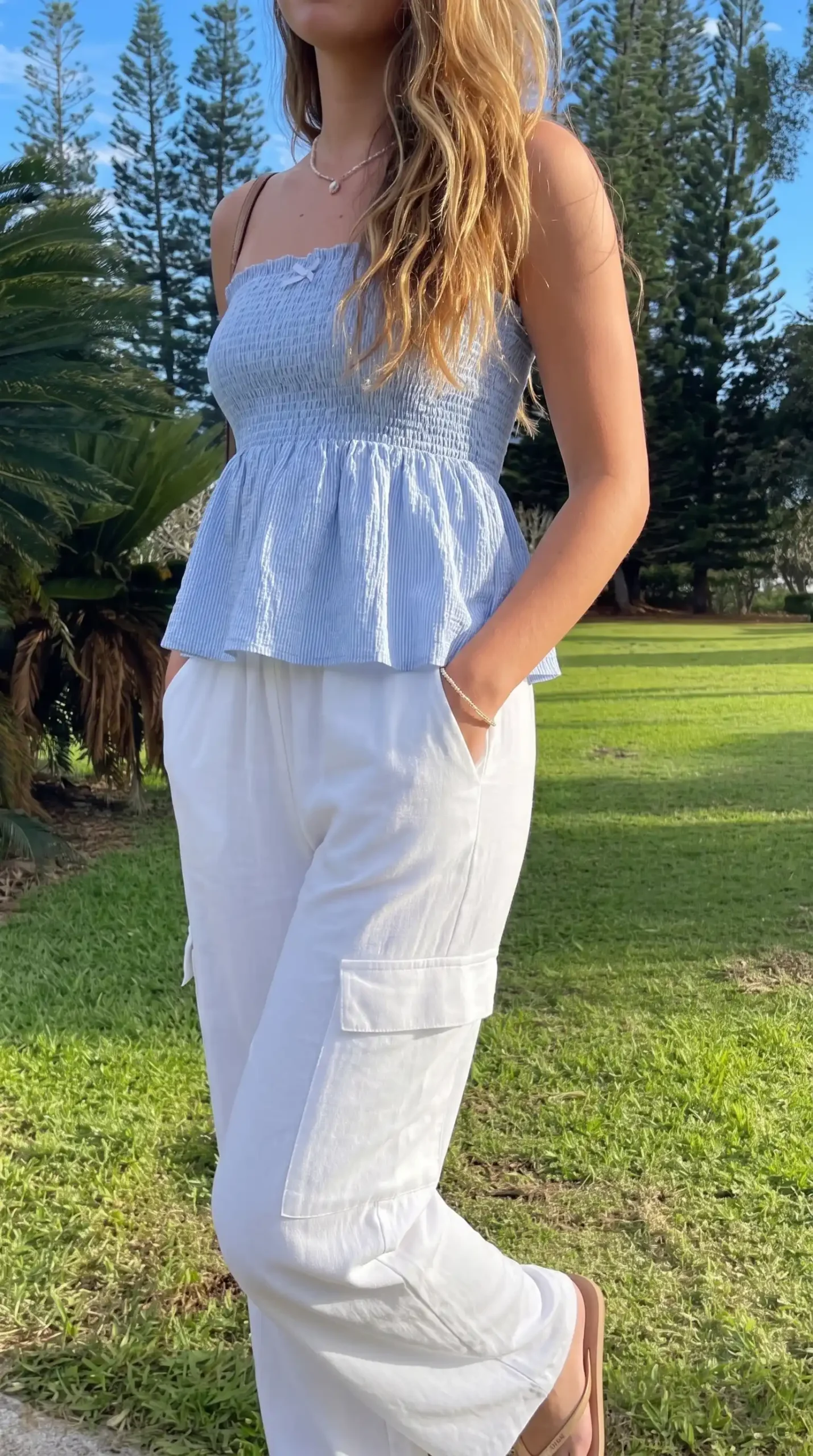 Woman wearing blue smocked peplum top with white wide-leg pants outdoors in park