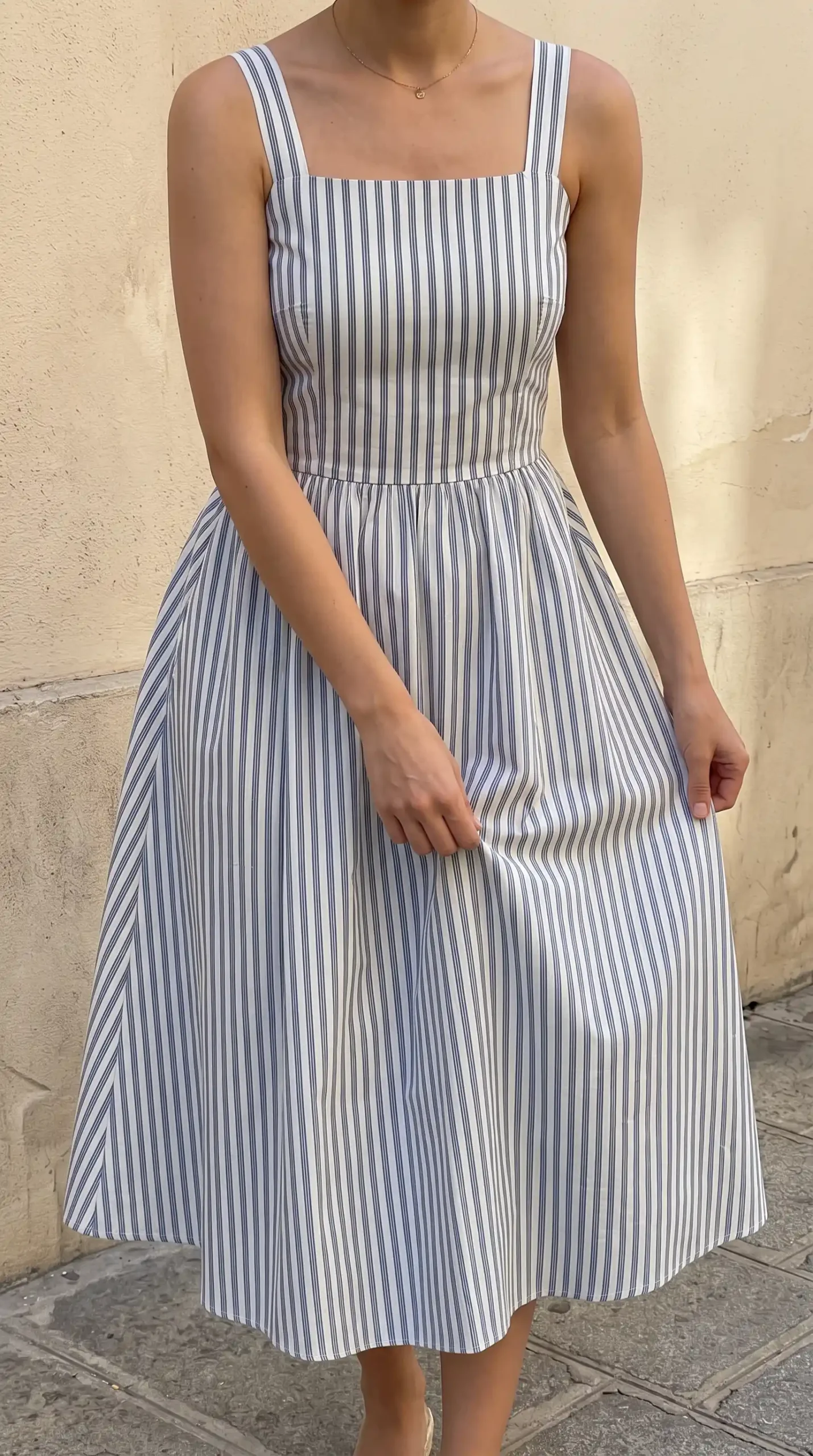 Woman wearing a blue and white striped dress with a straw handbag and white sandals outdoors near a red door
