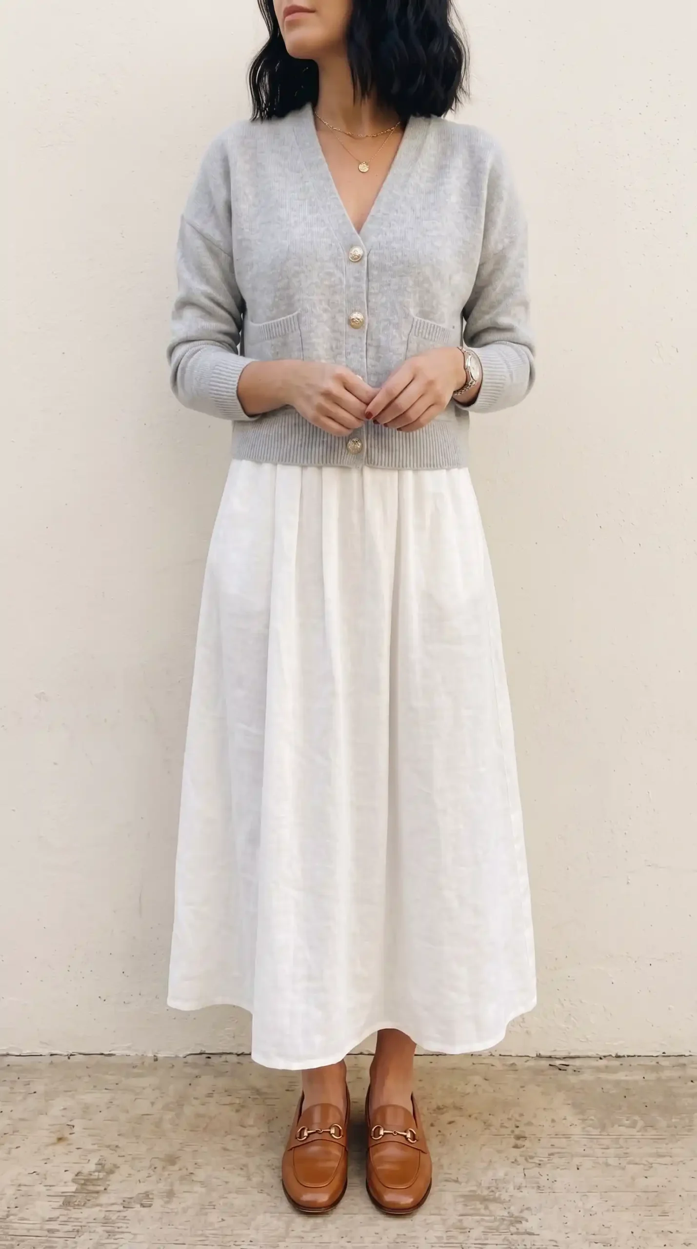 Woman wearing a light gray cardigan with a white skirt, beige loafers, and tan shoulder bag outdoors