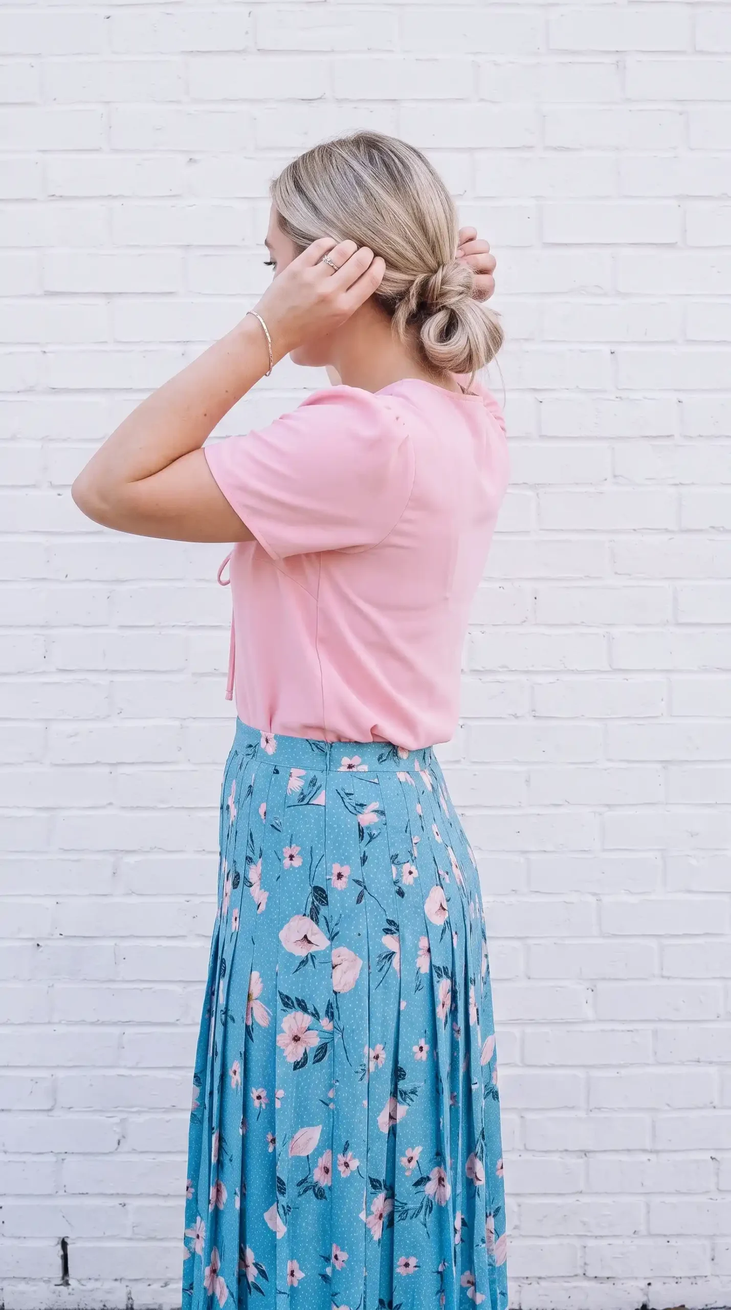 Woman wearing a light pink blouse with a blue floral pleated skirt, white espadrille wedges, and a straw handbag outdoors