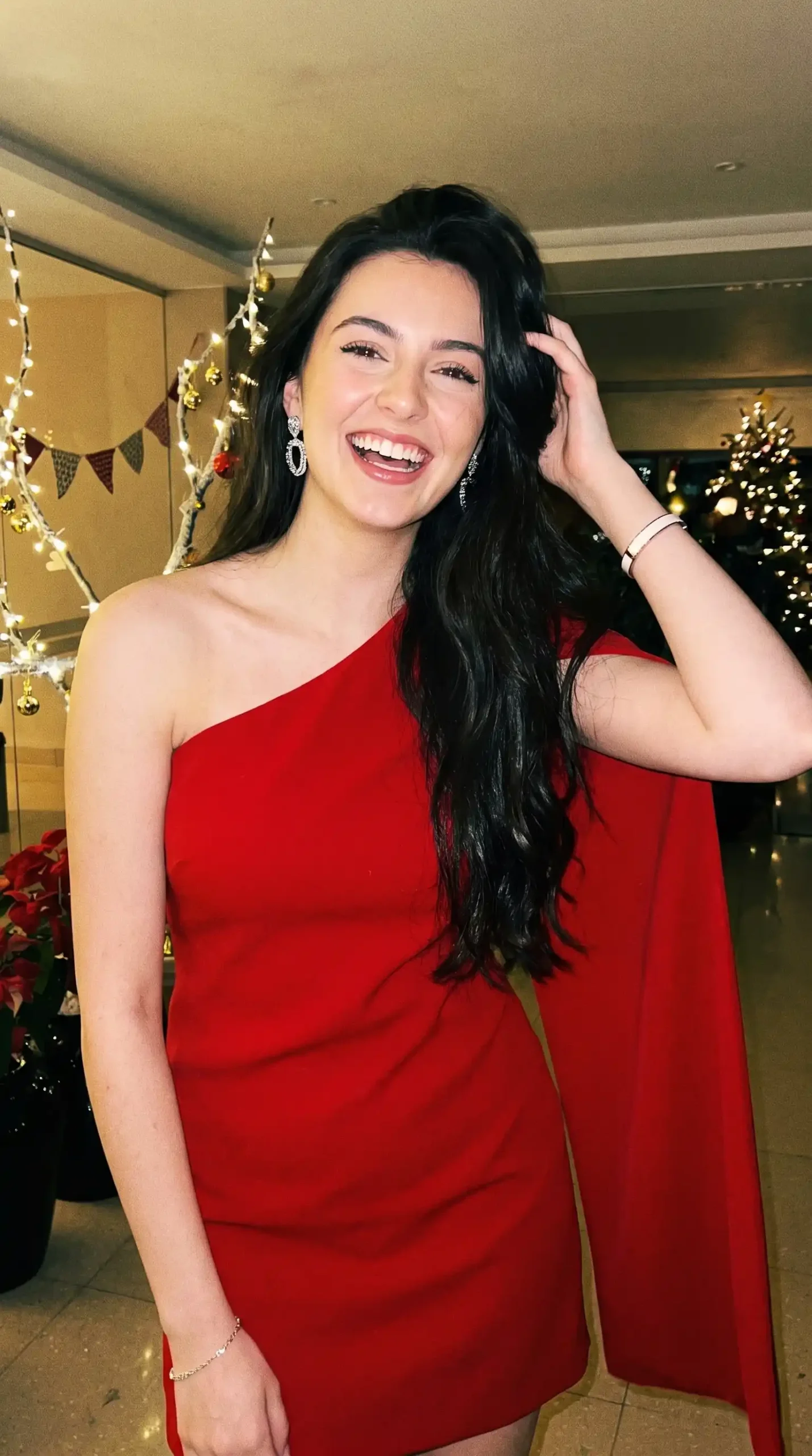 Woman wearing red one shoulder draped cape sleeve mini dress indoors at holiday party