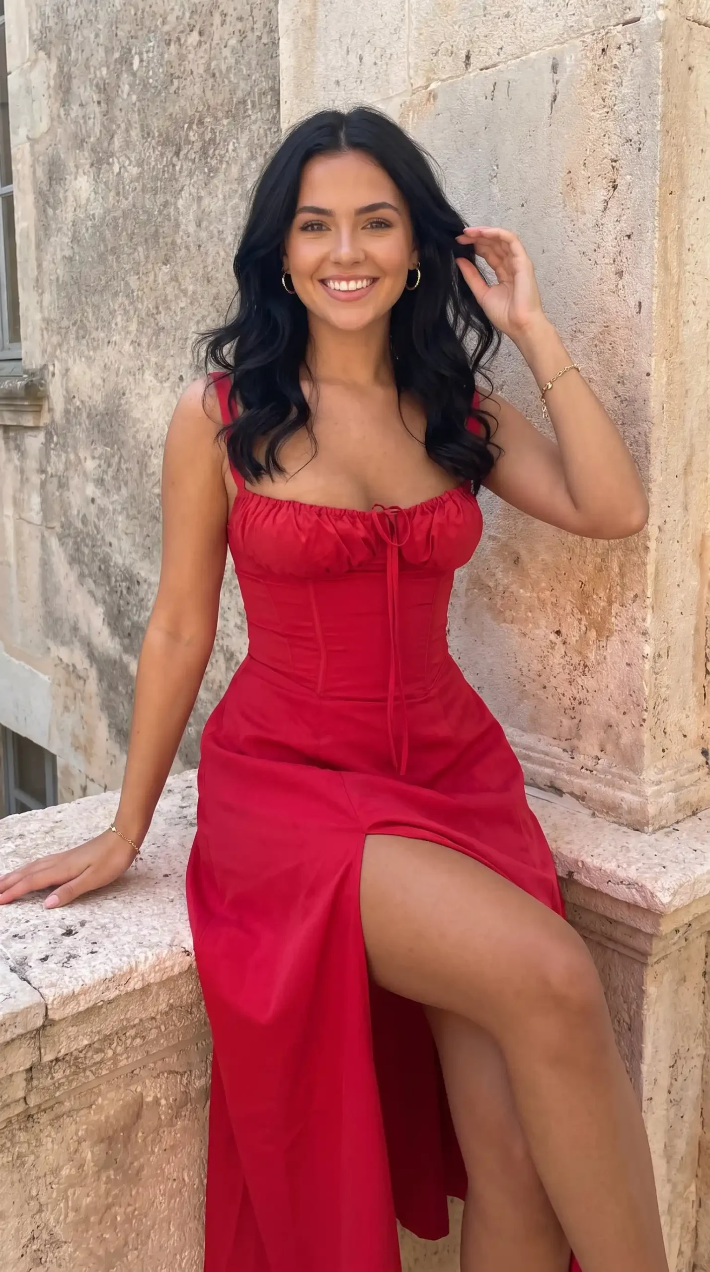 Woman wearing red tie front corset bodice high slit maxi dress sitting on stone ledge