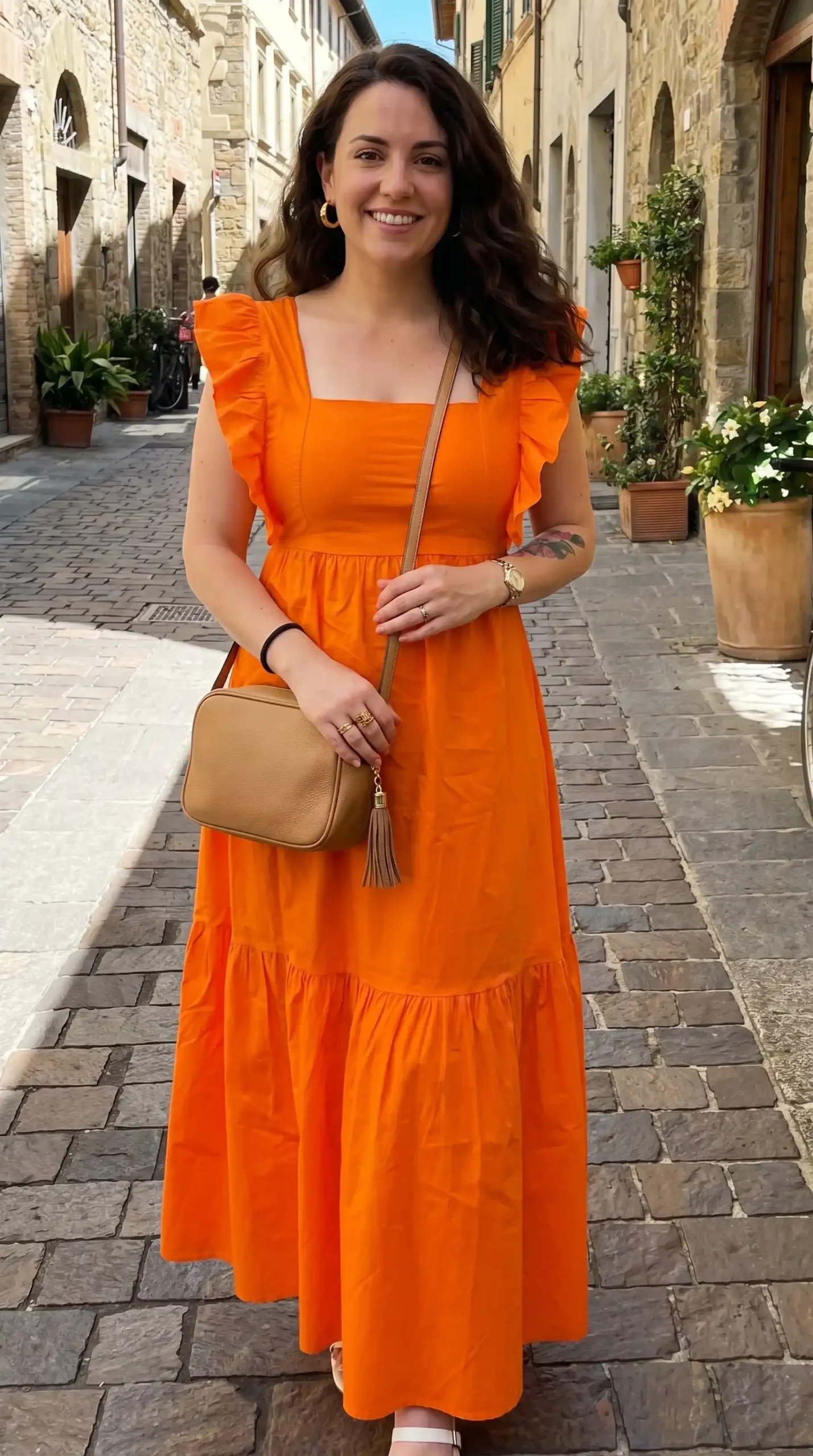 Woman wearing bright orange ruffle sleeve square neck tiered maxi dress with tan crossbody bag on cobblestone street