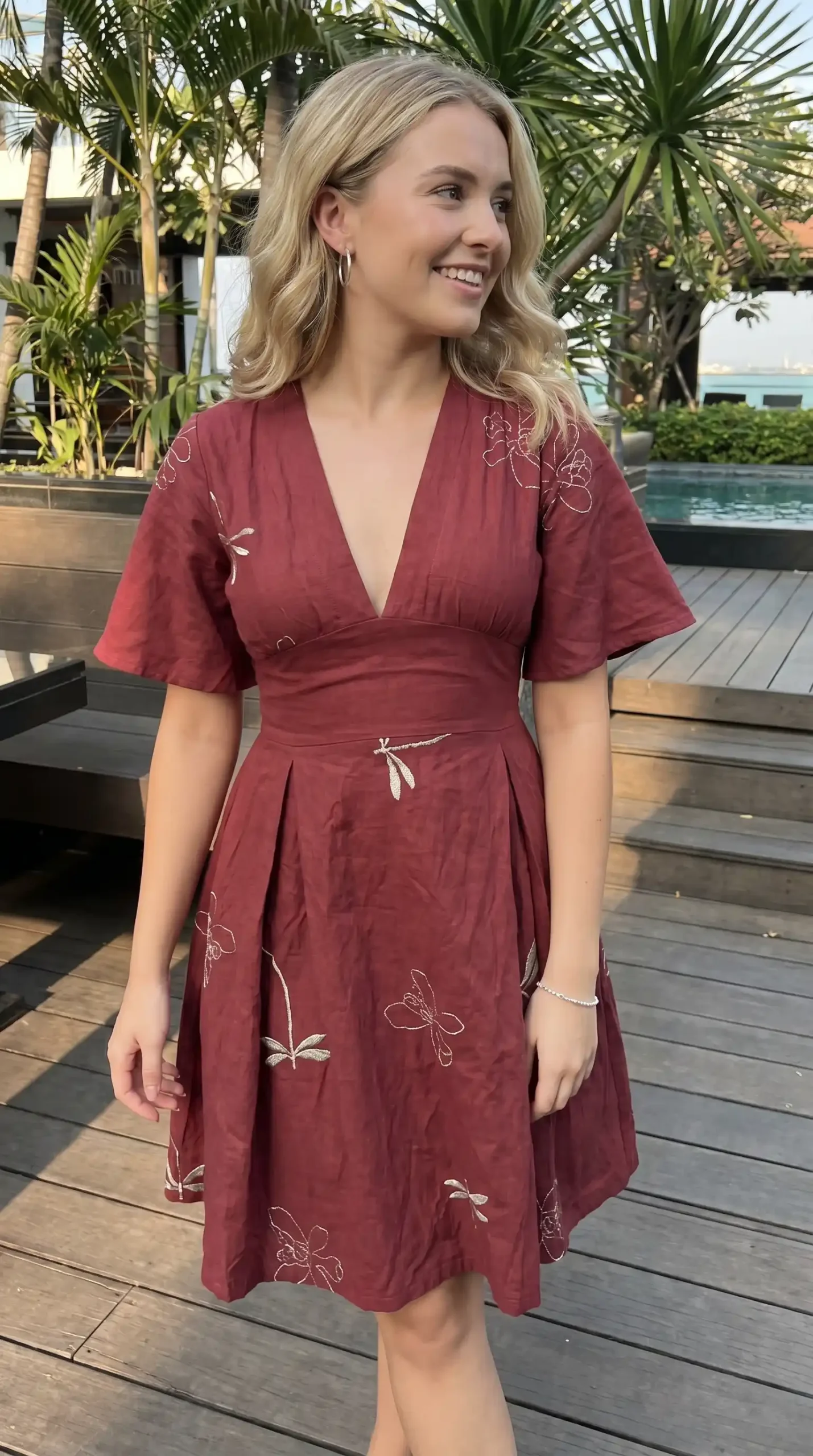 Woman wearing burgundy linen V neck embroidered flutter sleeve midi dress outdoors by pool in summer