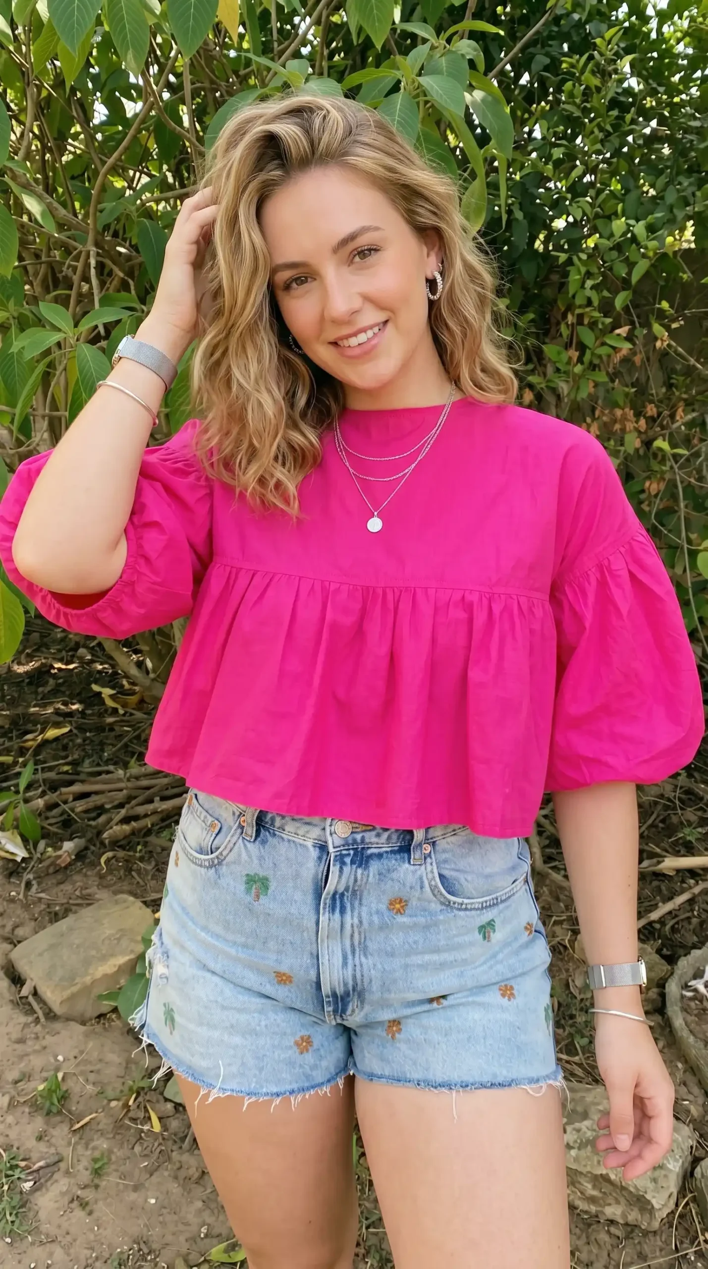 Woman wearing hot pink puff sleeve babydoll top with palm tree embroidered denim shorts outdoors