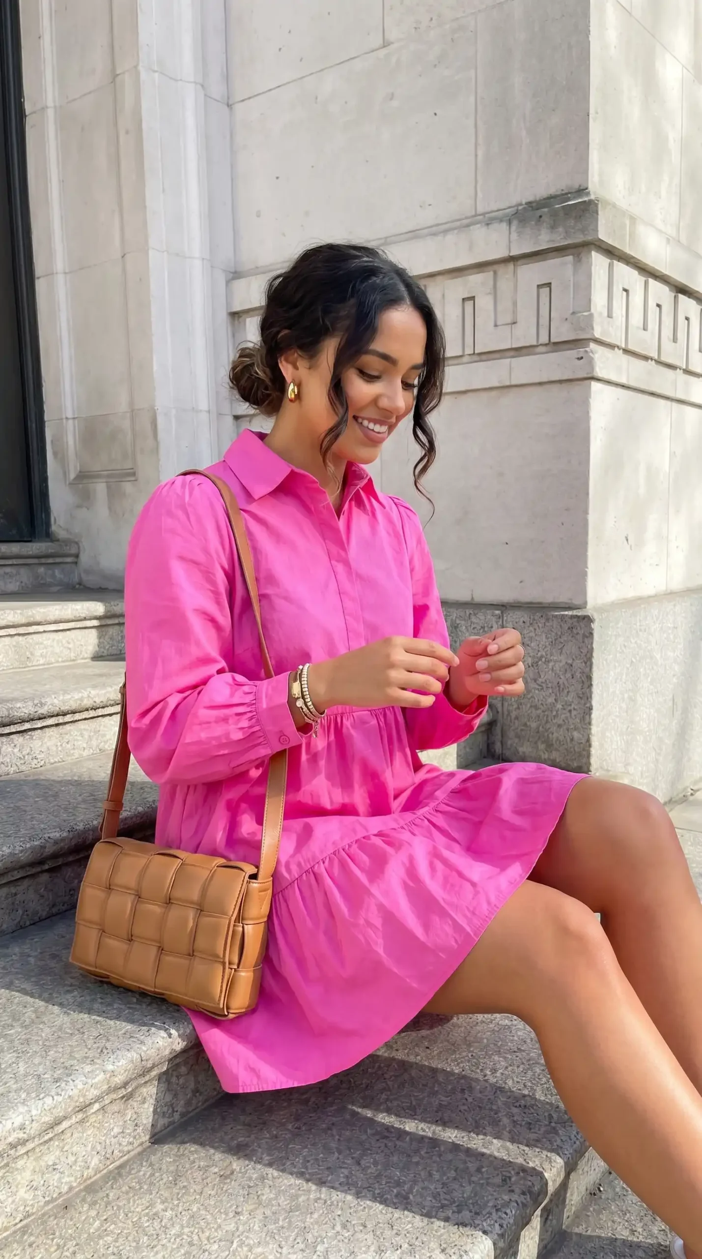 Woman wearing hot pink tiered shirt mini dress with tan crossbody bag sitting on stone steps