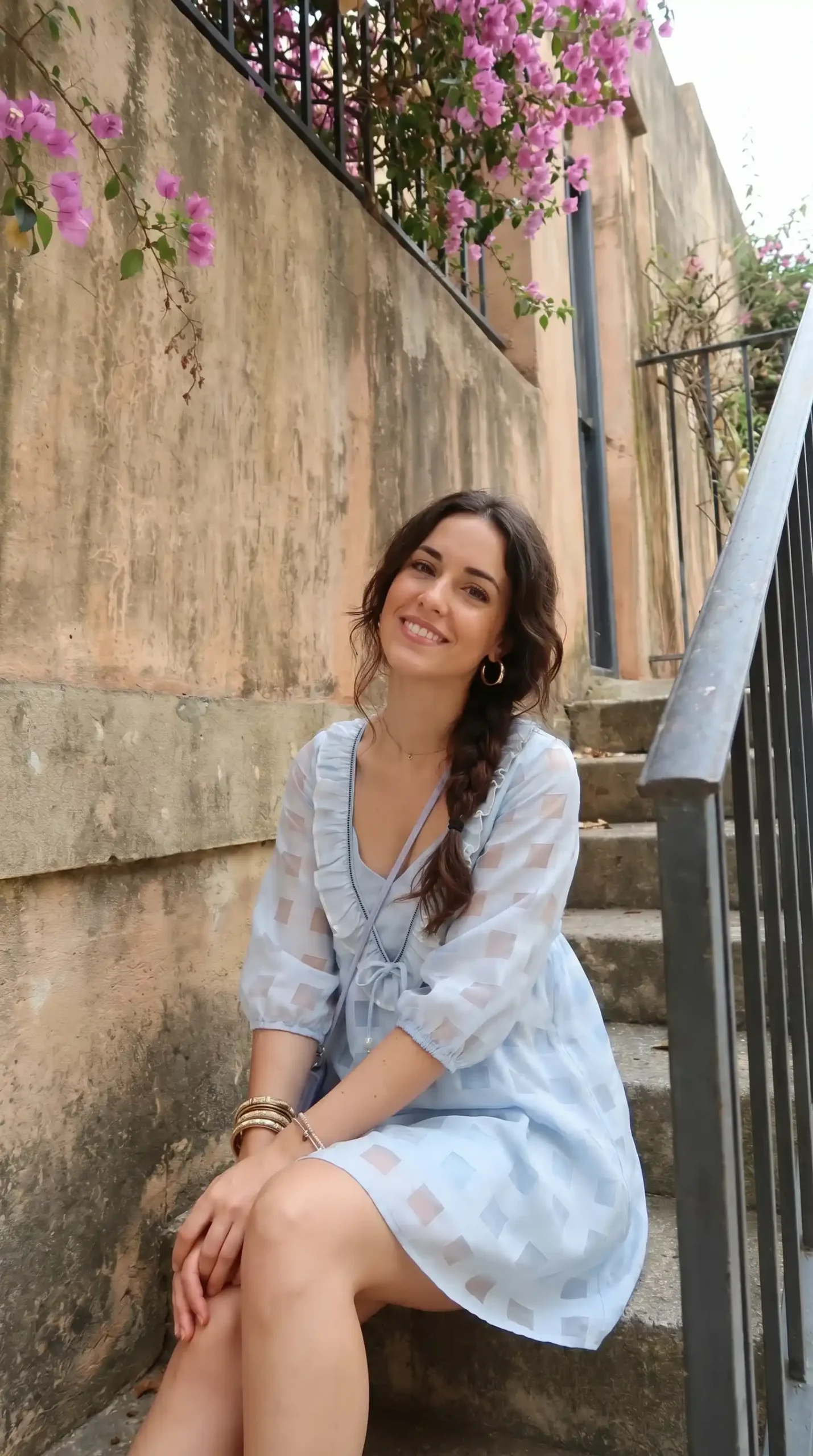 Woman wearing light blue checkered wrap dress with tie neckline in alley with flowers