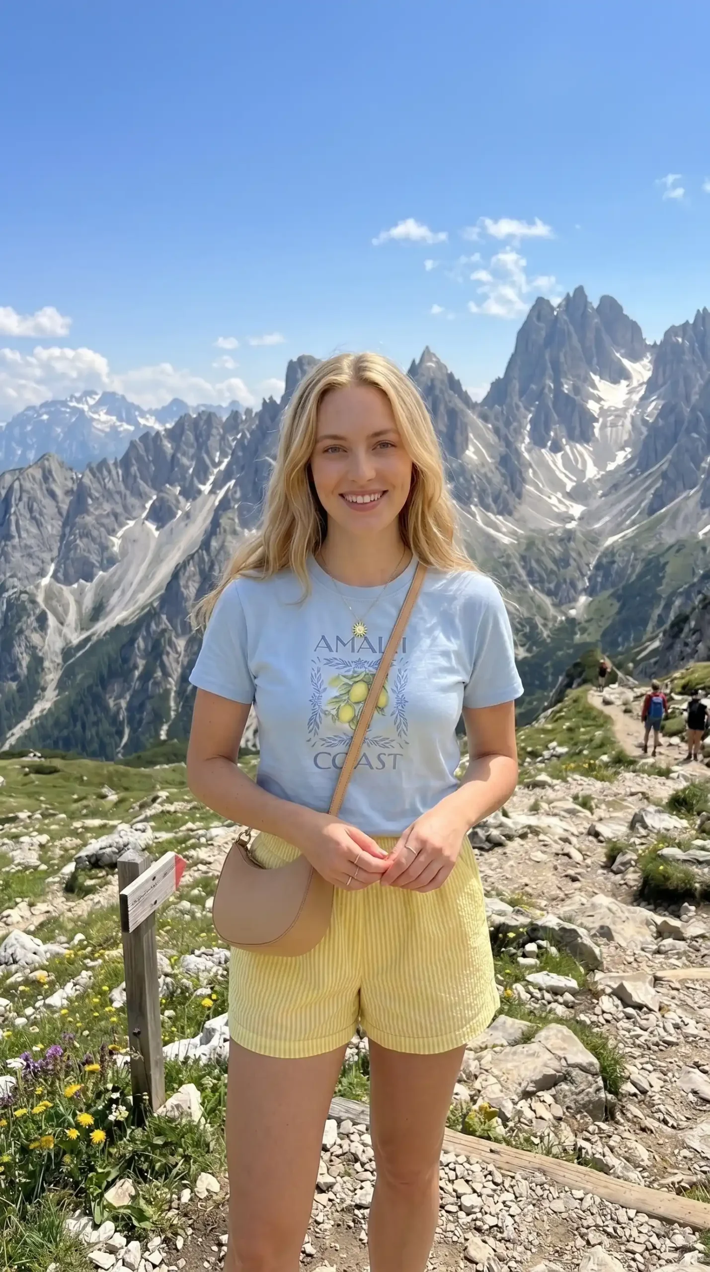 Woman wearing light blue Amalfi Coast graphic tee with yellow striped shorts and beige crossbody bag in mountains