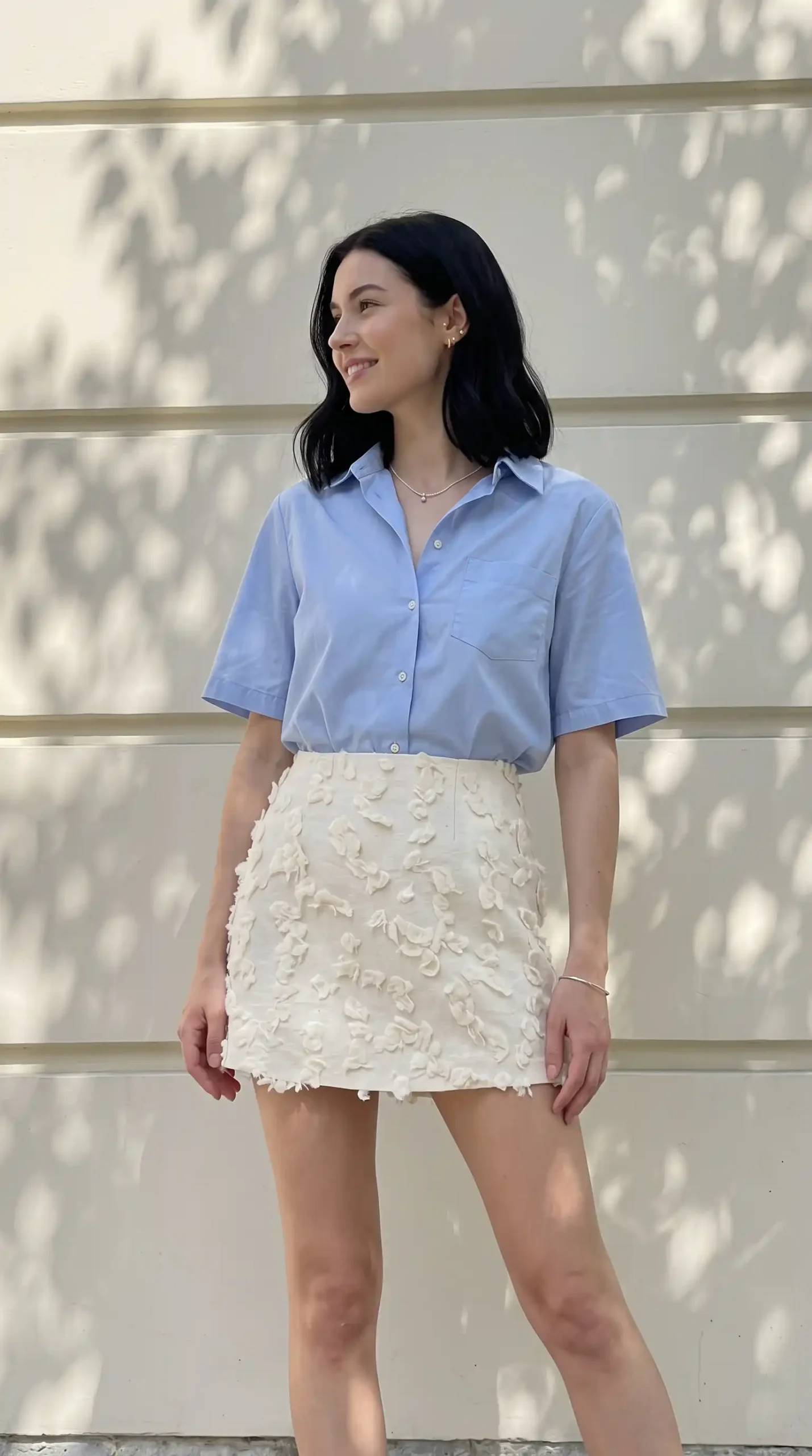 Woman wearing light blue short sleeve button up shirt with cream floral textured mini skirt outdoors