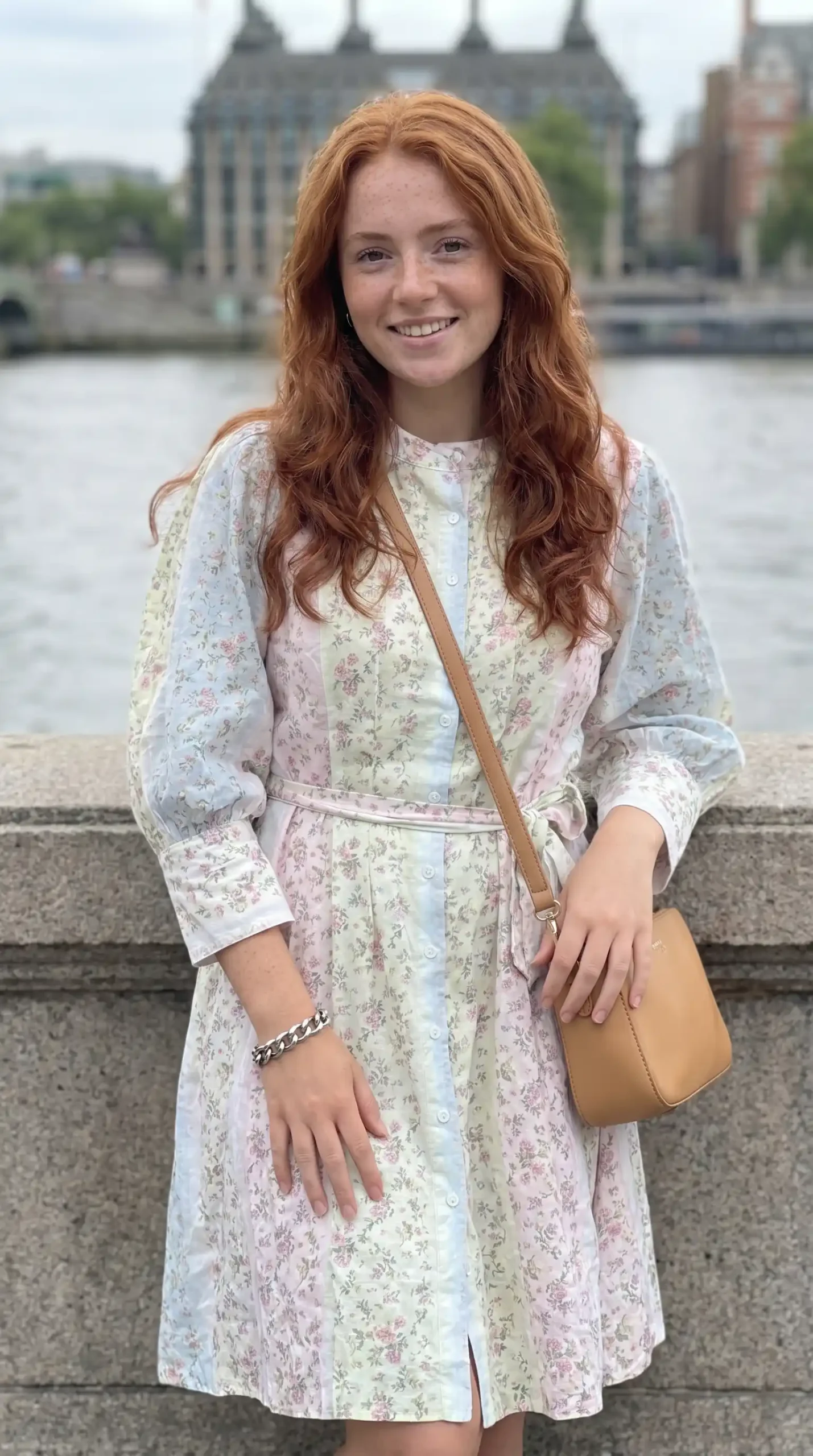 Woman wearing pastel multicolour floral patchwork button down midi dress with tan crossbody bag by riverside