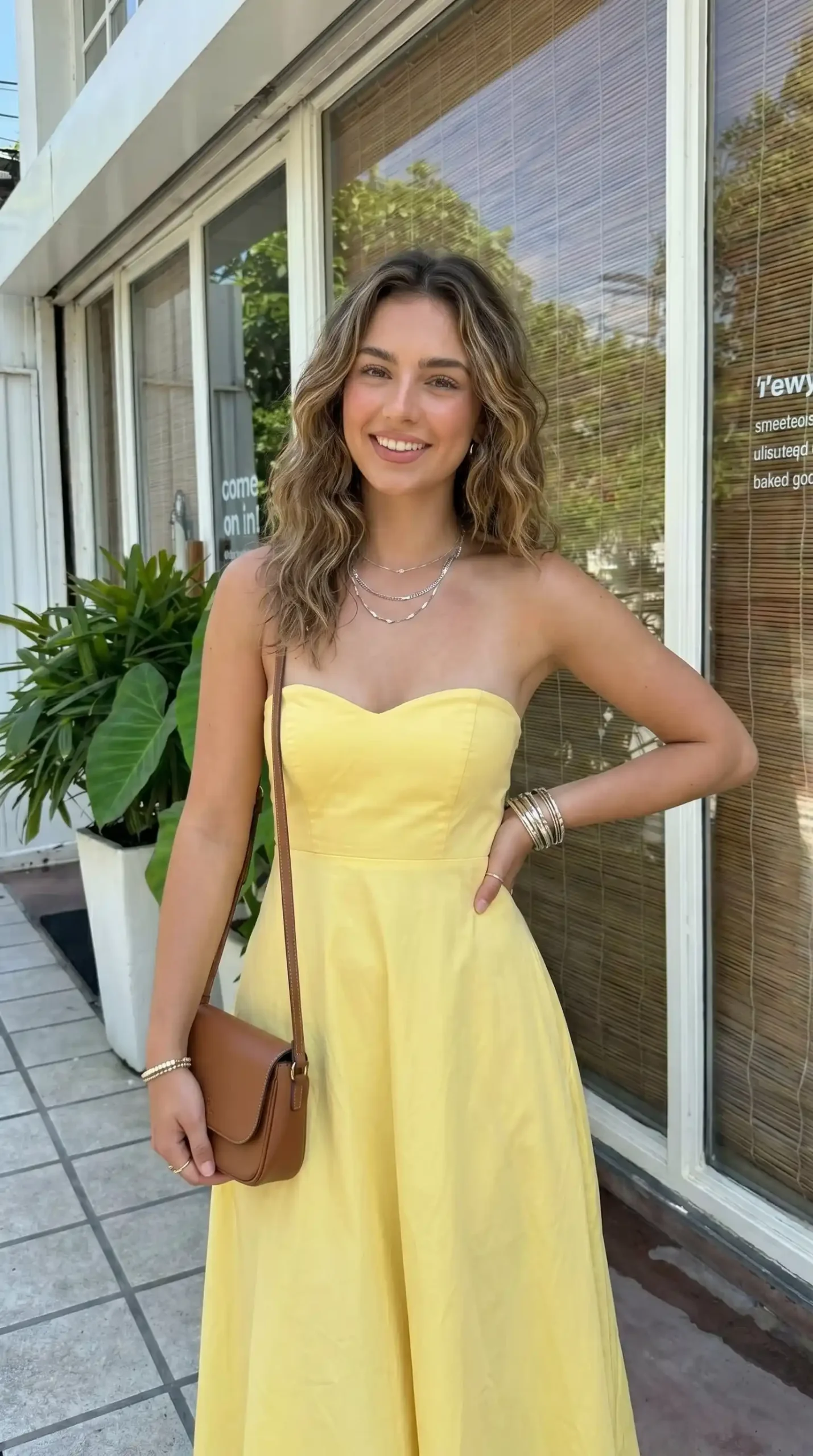 Woman wearing pastel yellow strapless sweetheart maxi dress with brown crossbody bag outdoors in summer