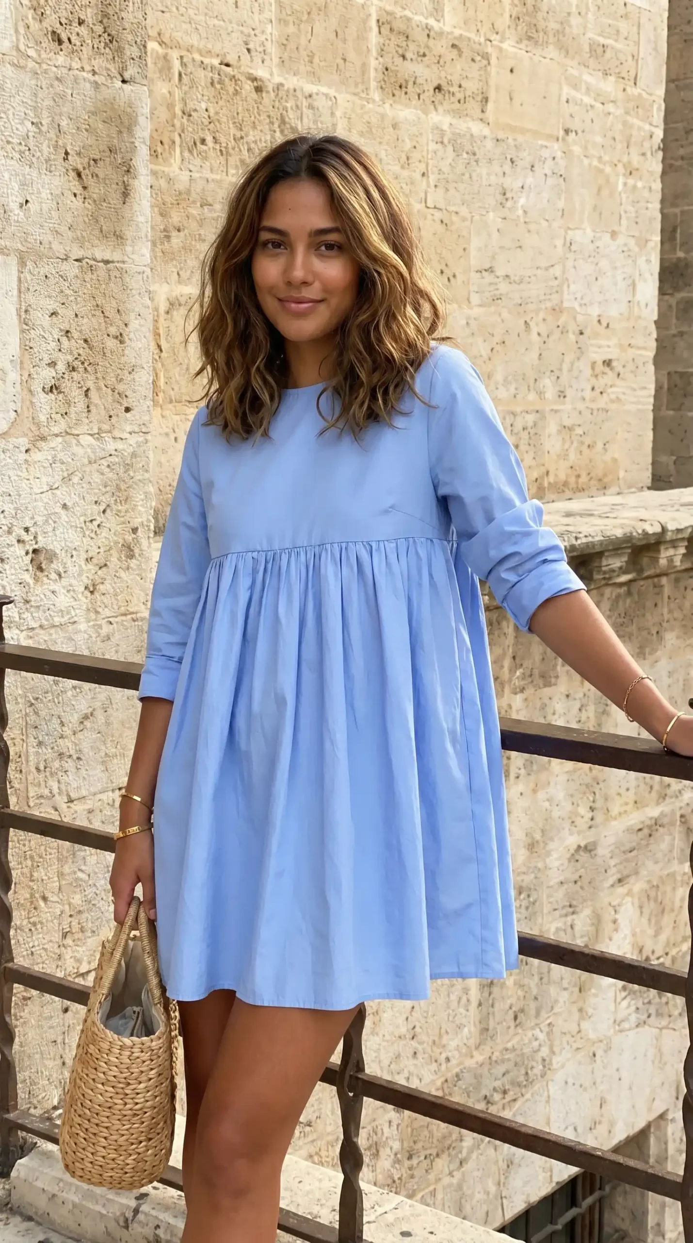Woman wearing periwinkle blue babydoll mini dress with woven straw bag by stone wall