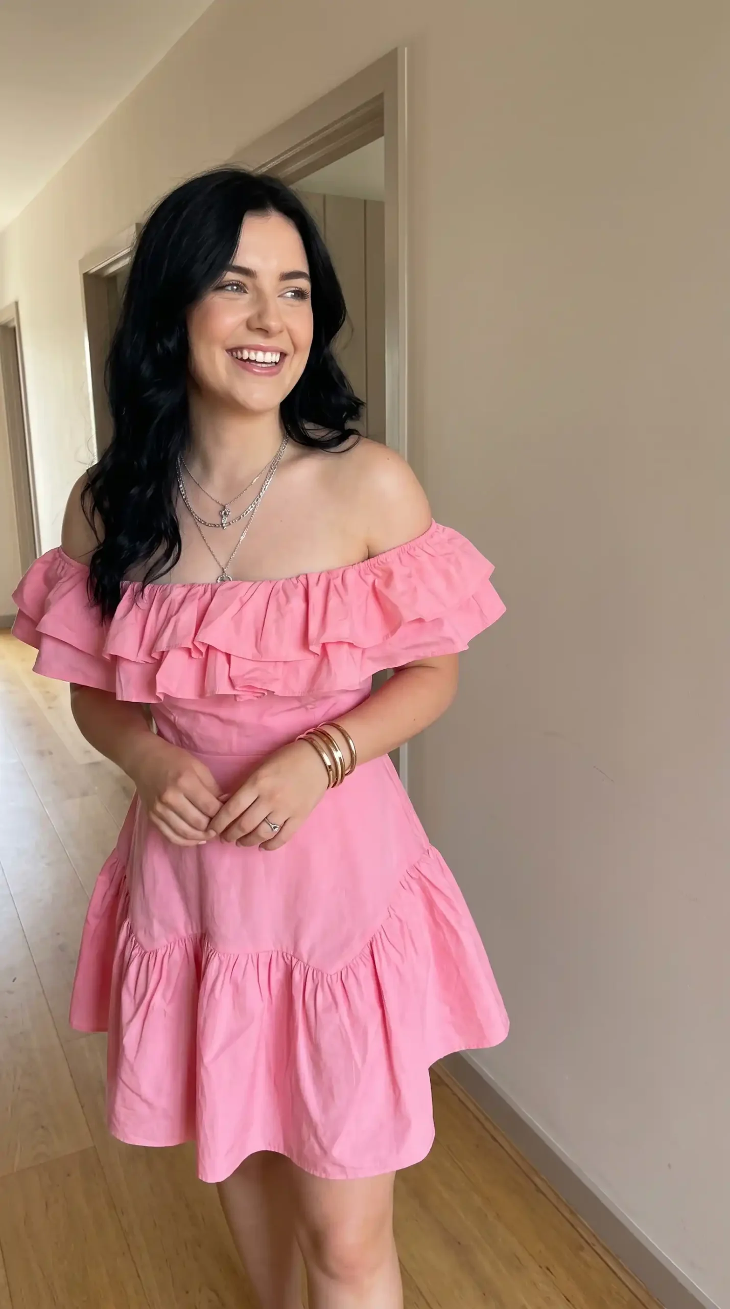 Woman wearing pink off shoulder ruffle tiered mini dress with gold bangles and layered necklaces indoors