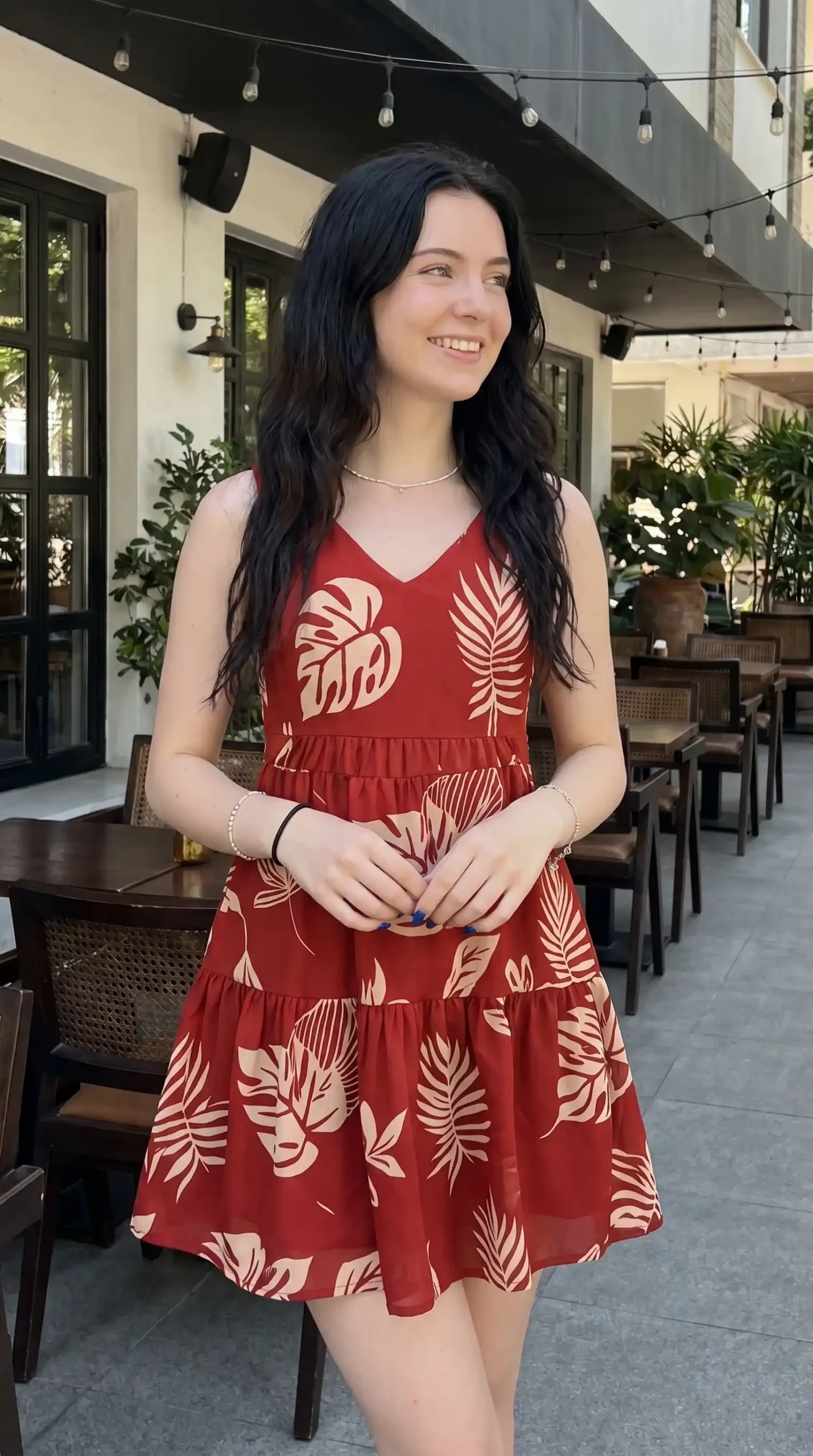Woman wearing rust red and cream tropical leaf print V neck tiered mini dress at outdoor cafe in summer