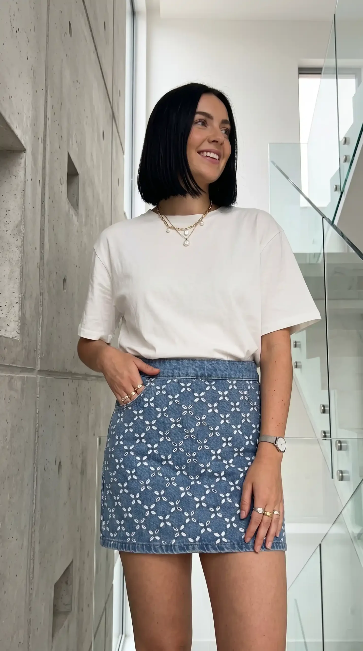 Woman wearing white oversized tee with blue denim floral embroidered mini skirt and gold layered necklaces indoors