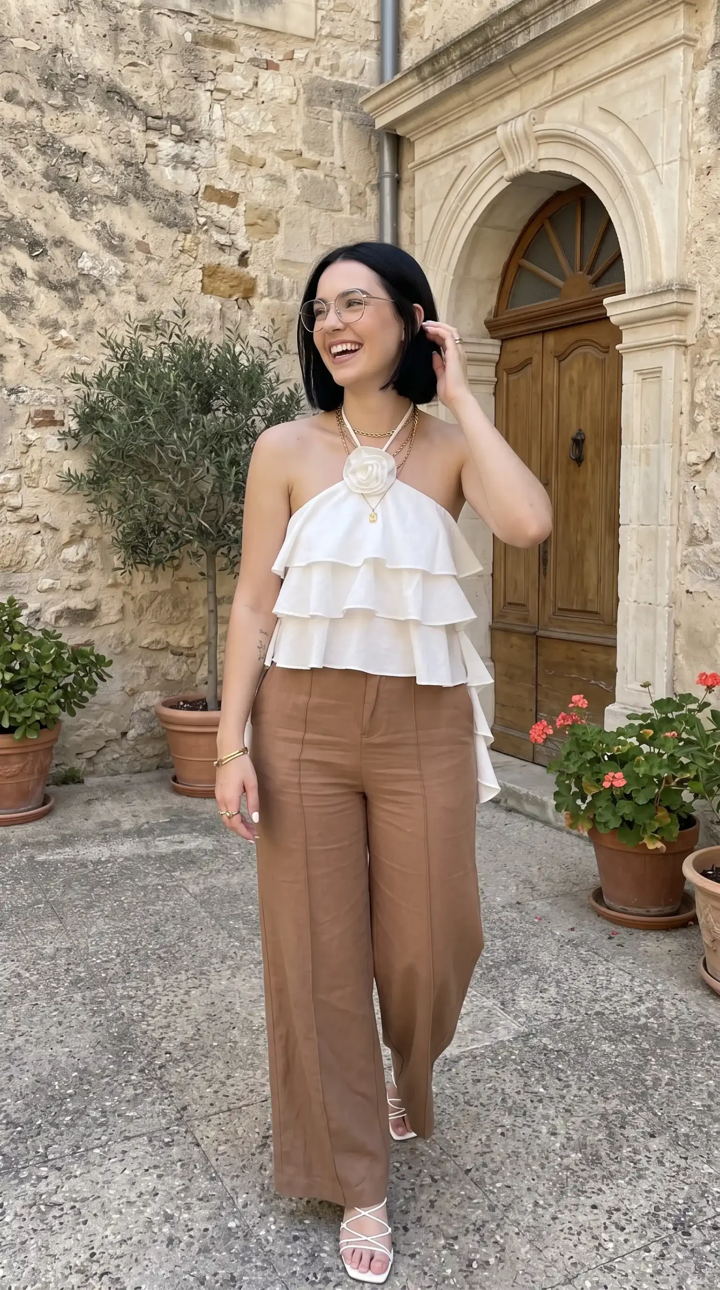 Woman wearing white tiered ruffle rosette halter top with brown wide leg linen pants and white strappy heels outdoors
