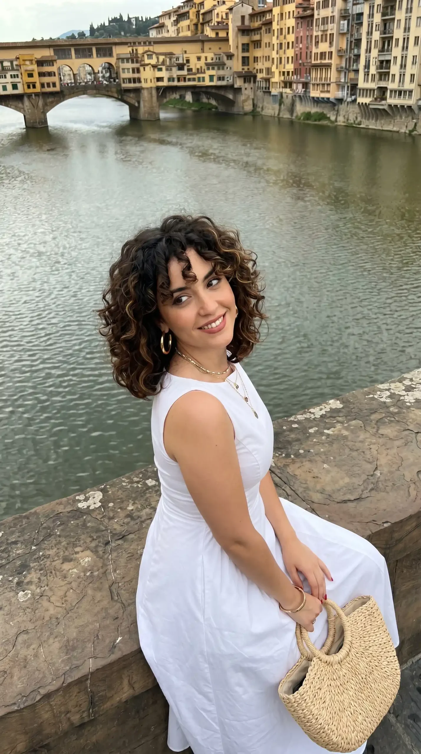 Woman wearing white sleeveless midi dress with straw bag by riverside bridge