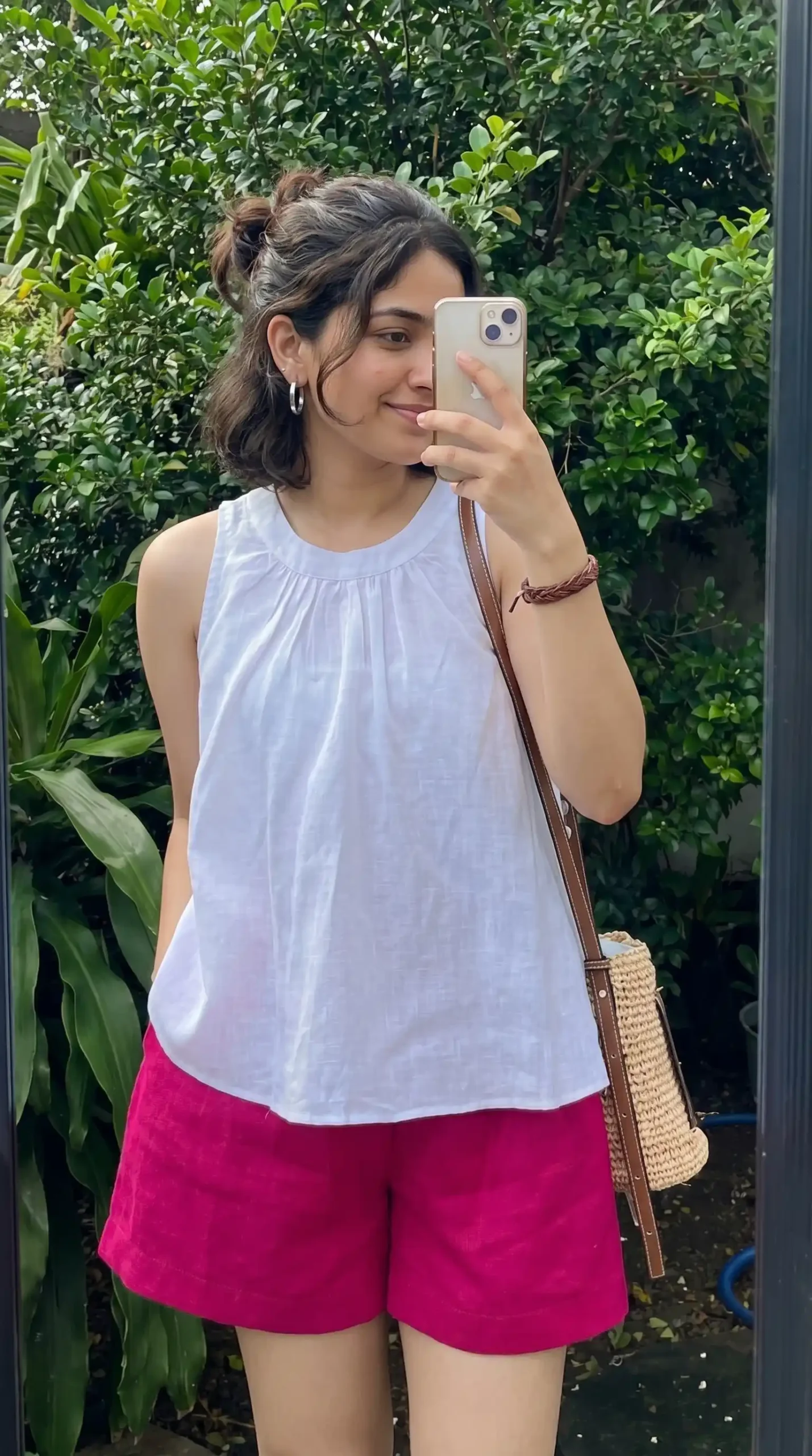 Woman wearing white sleeveless tank top with hot pink shorts and woven crossbody bag in garden