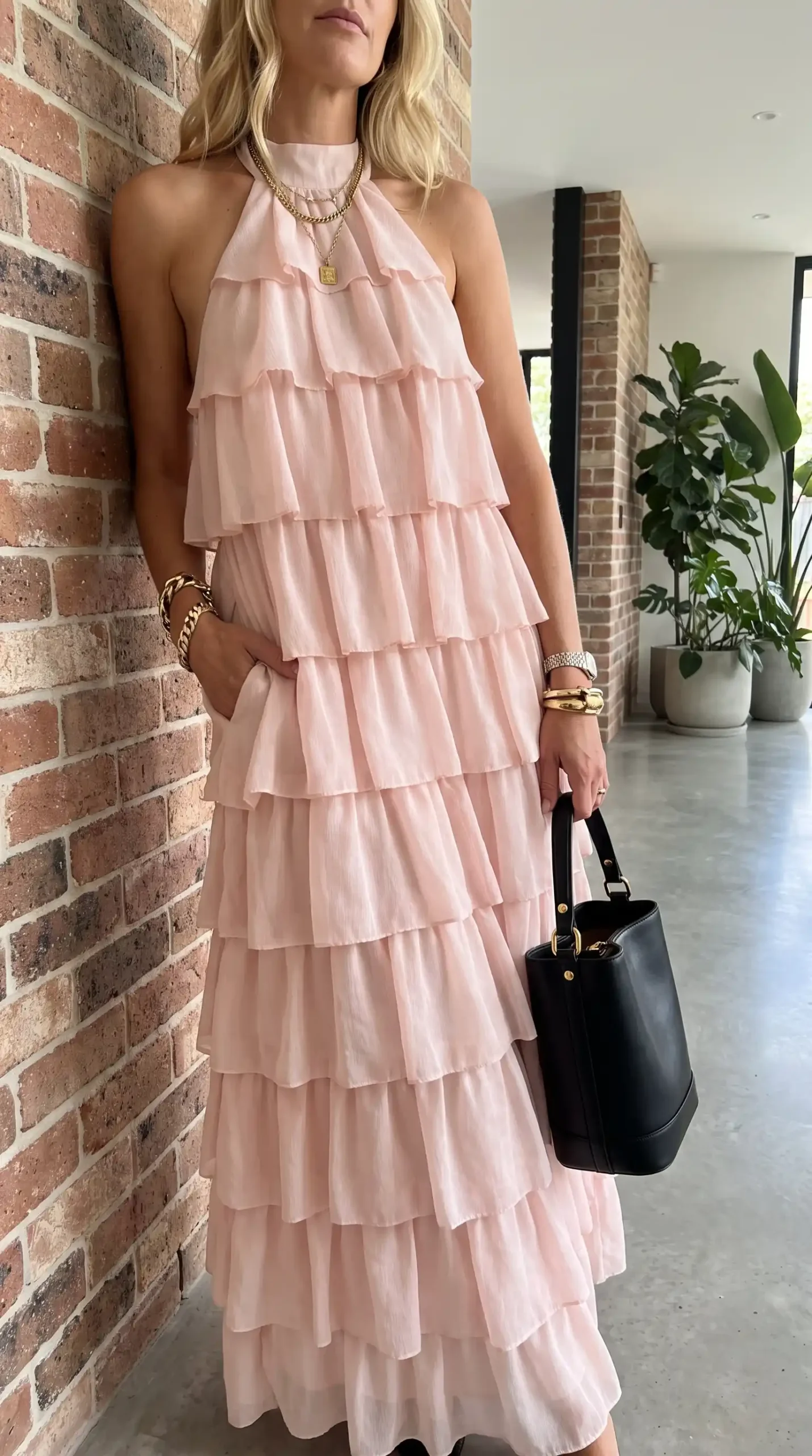 Woman wearing blush pink halter neck tiered ruffle maxi dress with black bag and gold jewelry