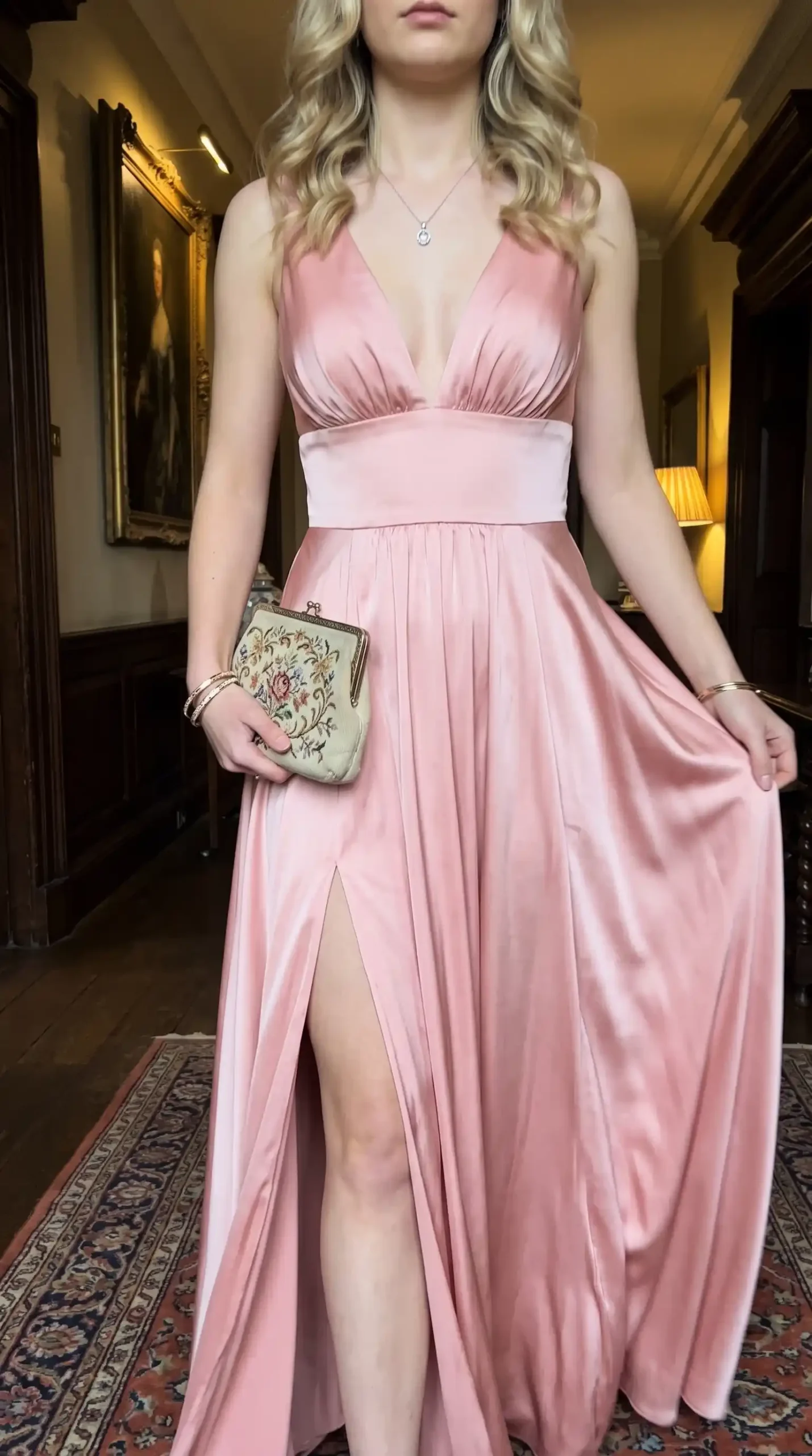 Woman wearing blush pink satin deep v neck thigh slit maxi dress holding embroidered clutch