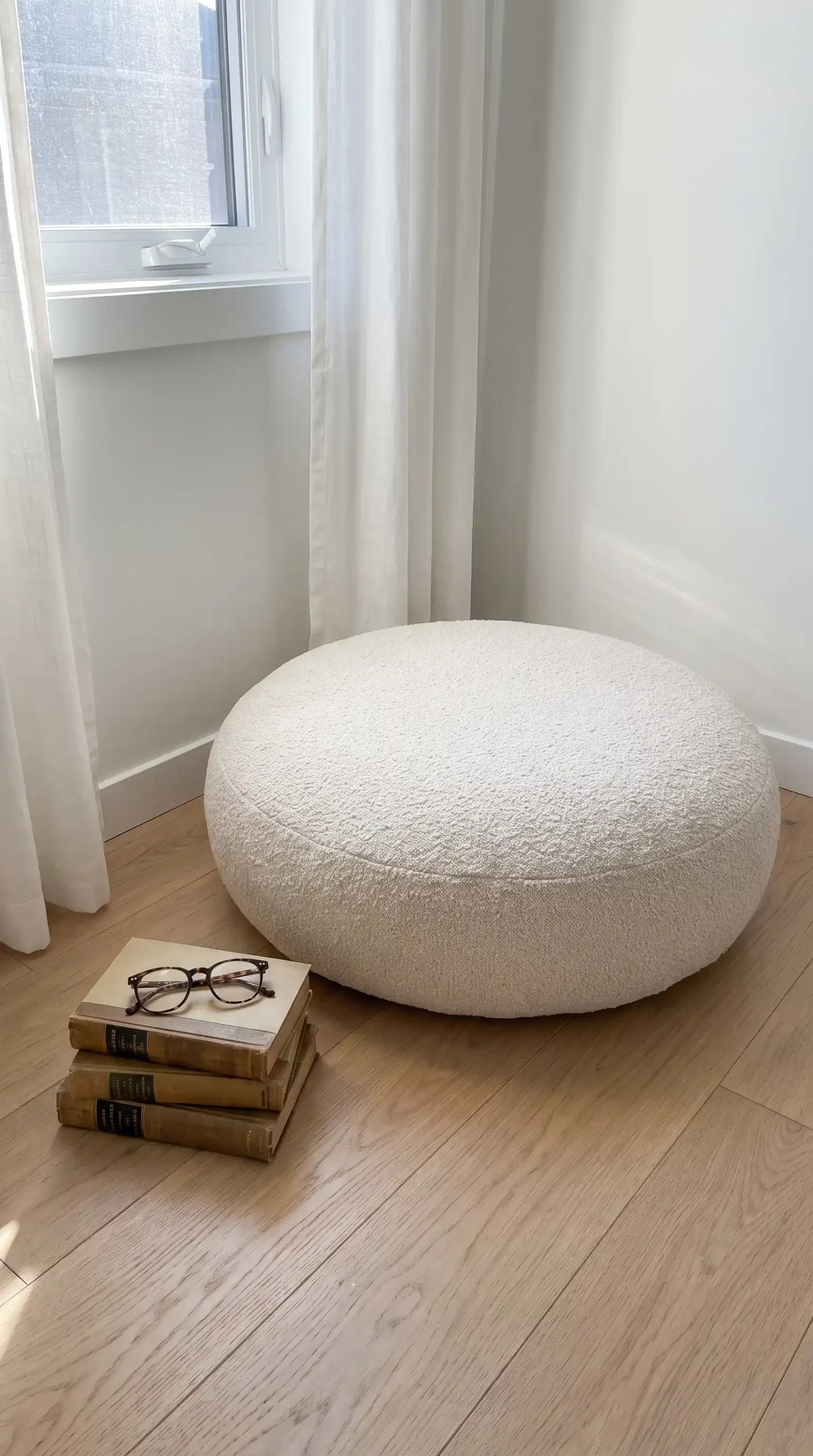 Large round cream boucle floor cushion beside stack of books on light oak bedroom floor
