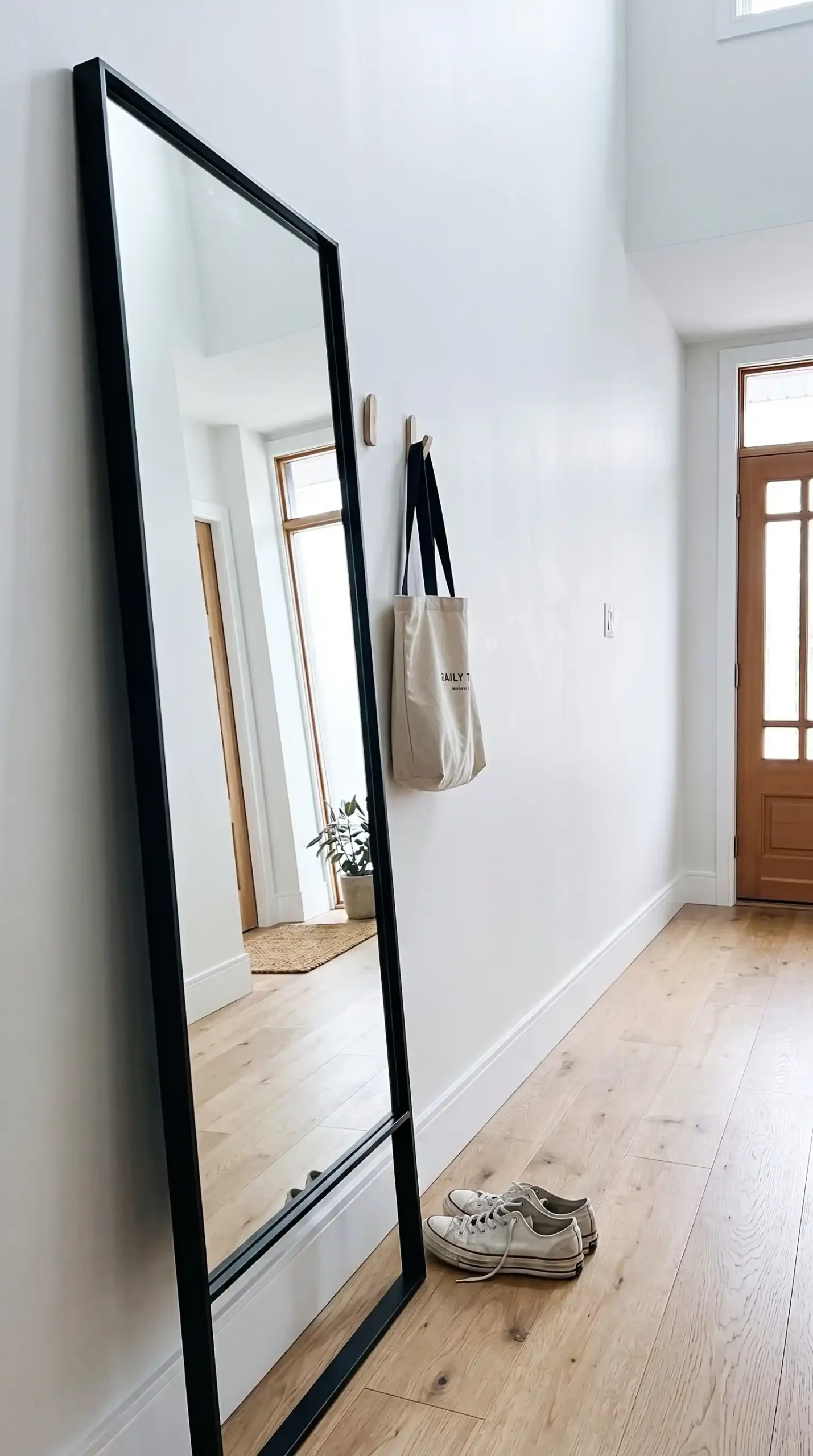 Very tall narrow black framed mirror leaning against white hallway wall beside wooden bench with sneakers below
