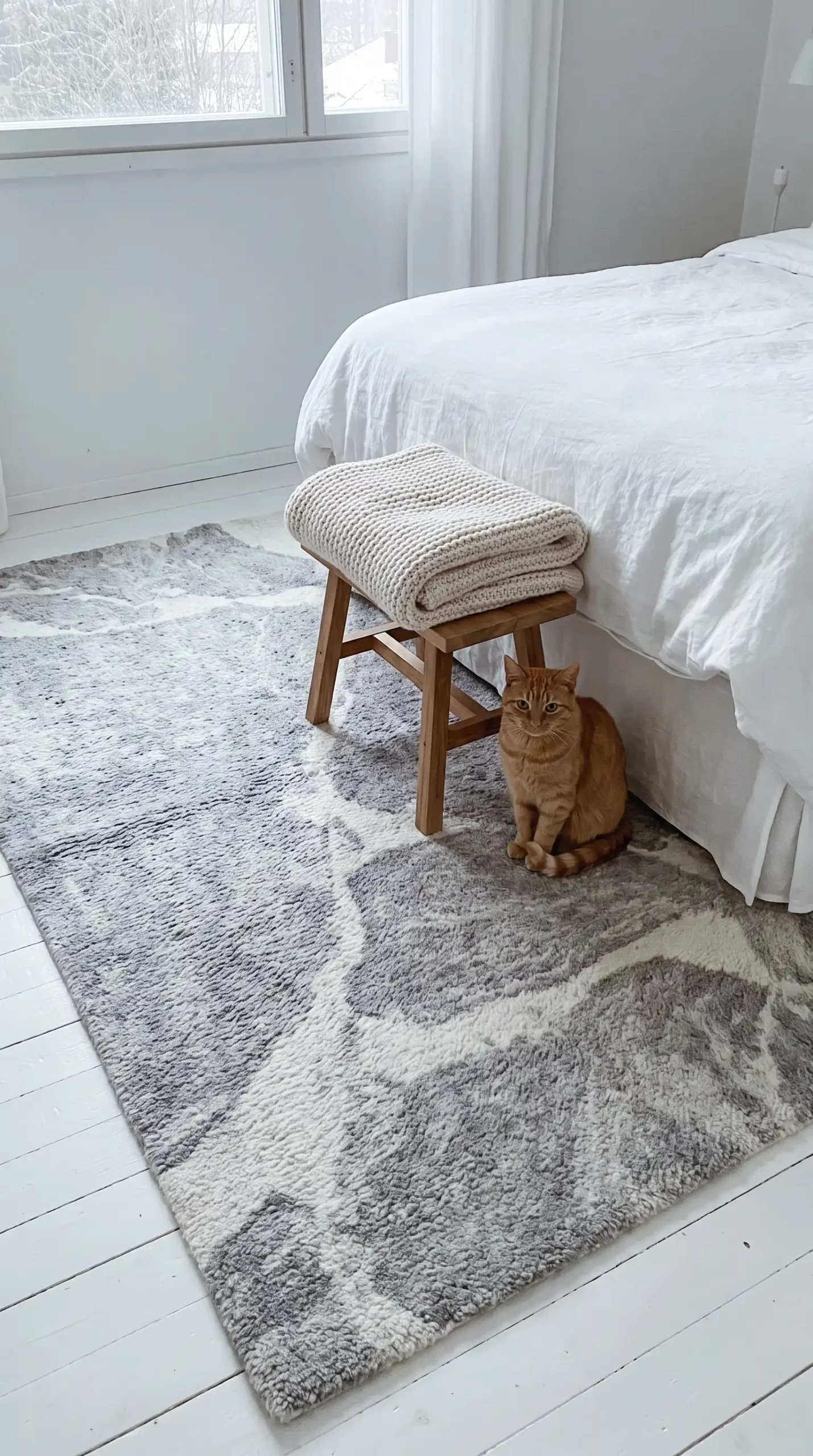 Large soft grey and white abstract pattern wool rug on white painted Scandinavian bedroom floor with wooden stool and knit blanket