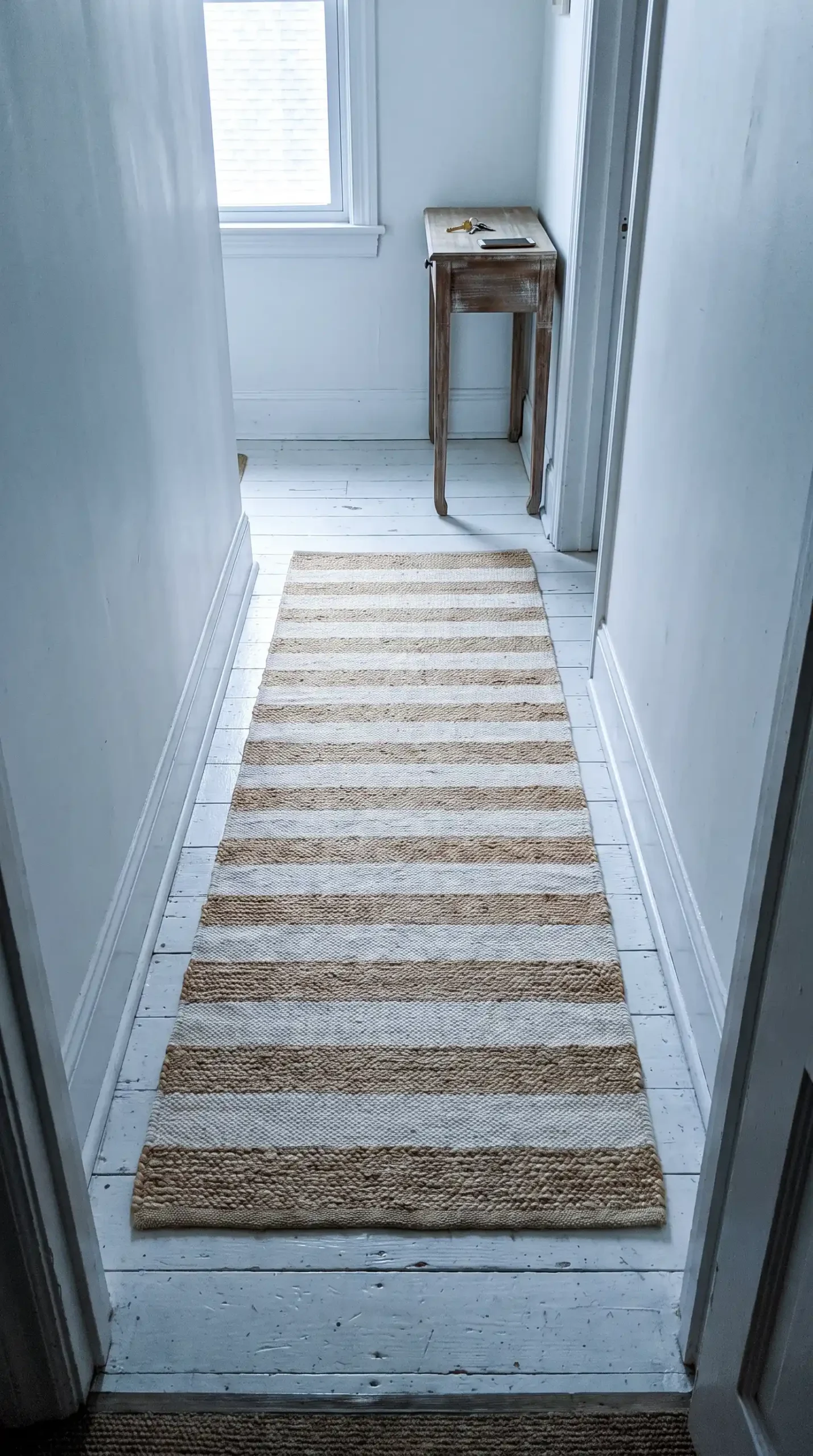 Long narrow cream and jute striped runner rug on white painted wood hallway floor with console table at end