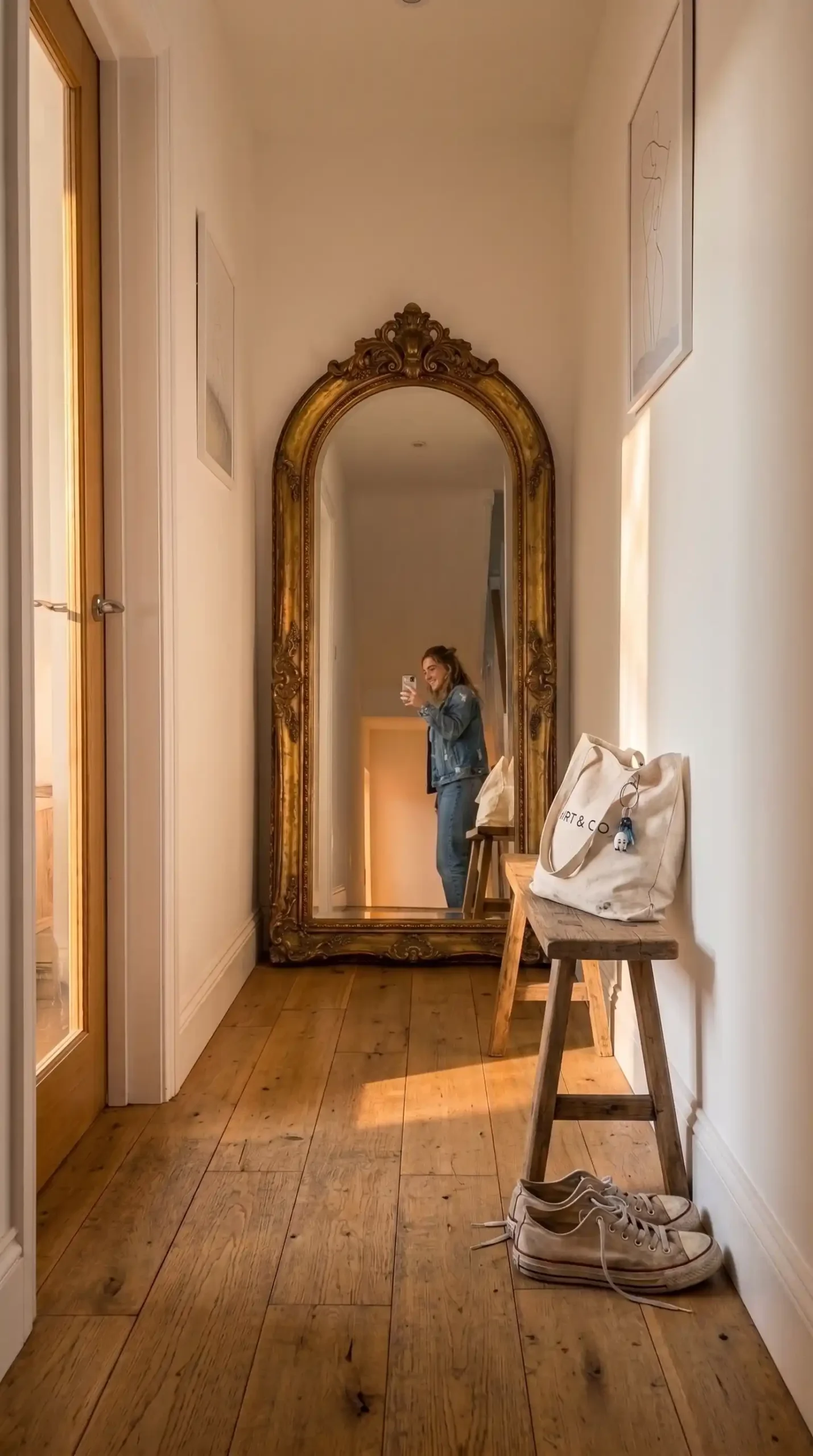 Large oversized vintage gold arch mirror leaning against white wall at end of hallway with wooden bench