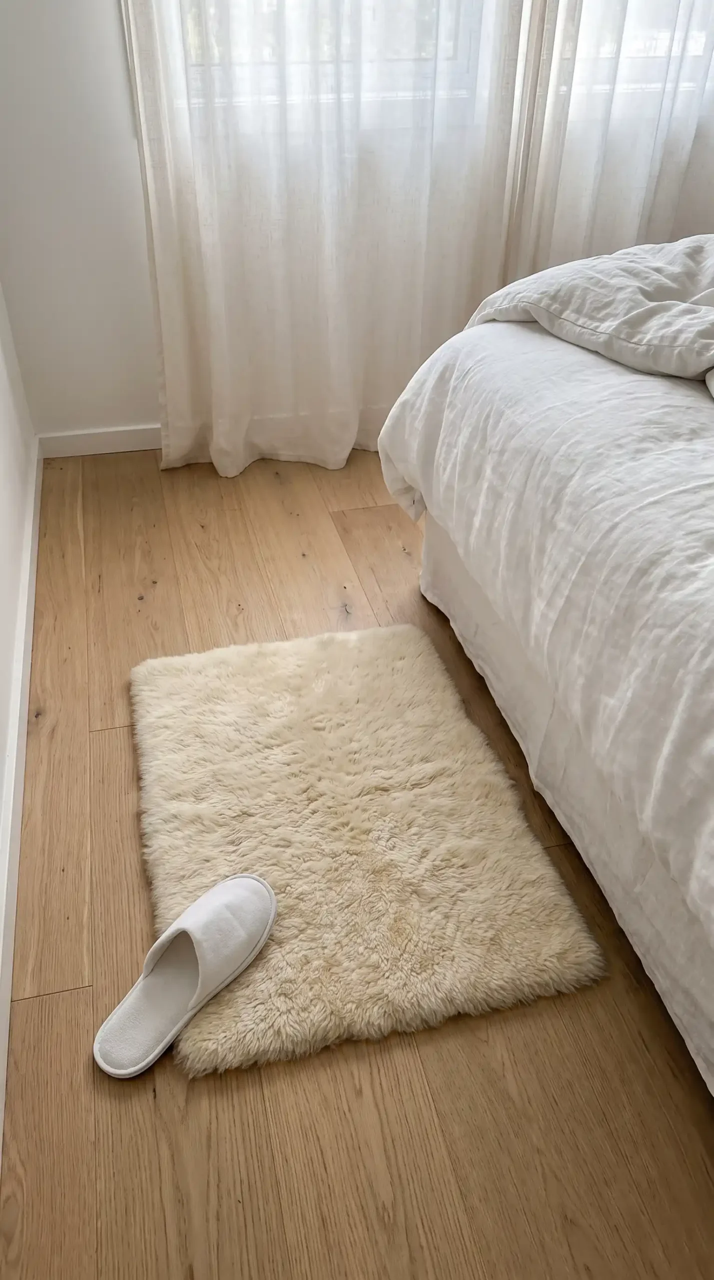 Small square cream sheepskin rug beside bed on light oak minimal bedroom floor with single slipper